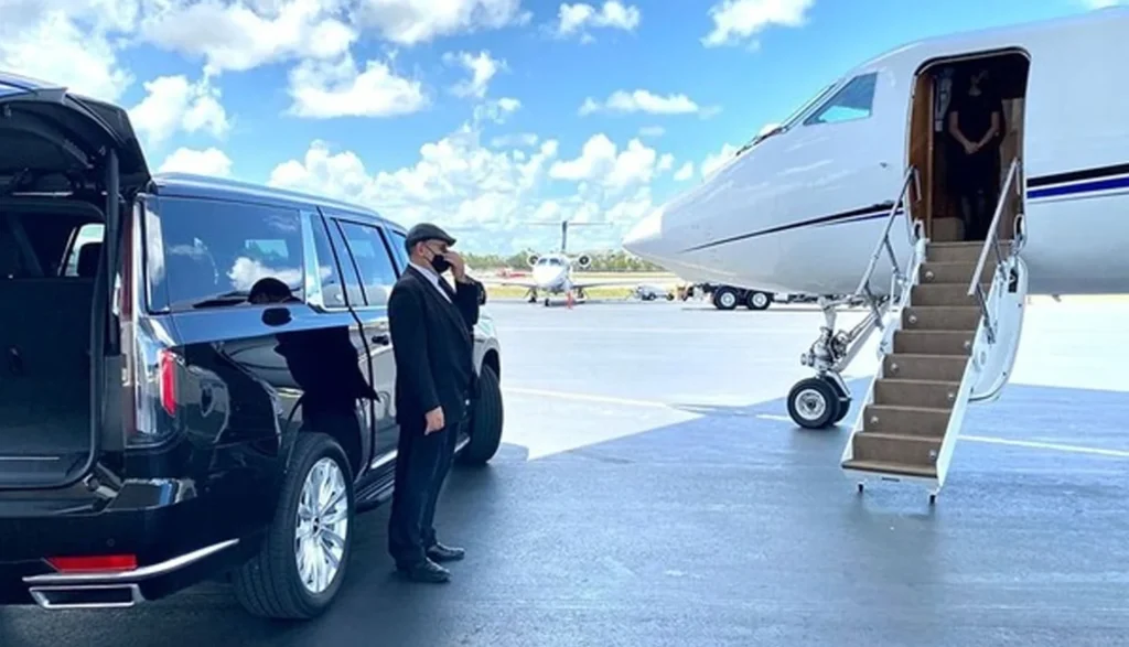 Man in a suit stands beside a black limousine, with a private jet visible in the background at a Los Angeles airport